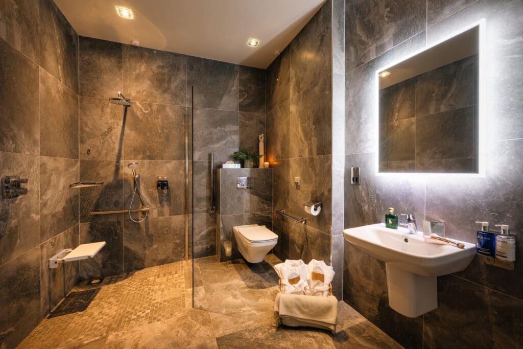 Accessible bathroom with level-access shower, support rails, wall-mounted sink and illuminated mirror.