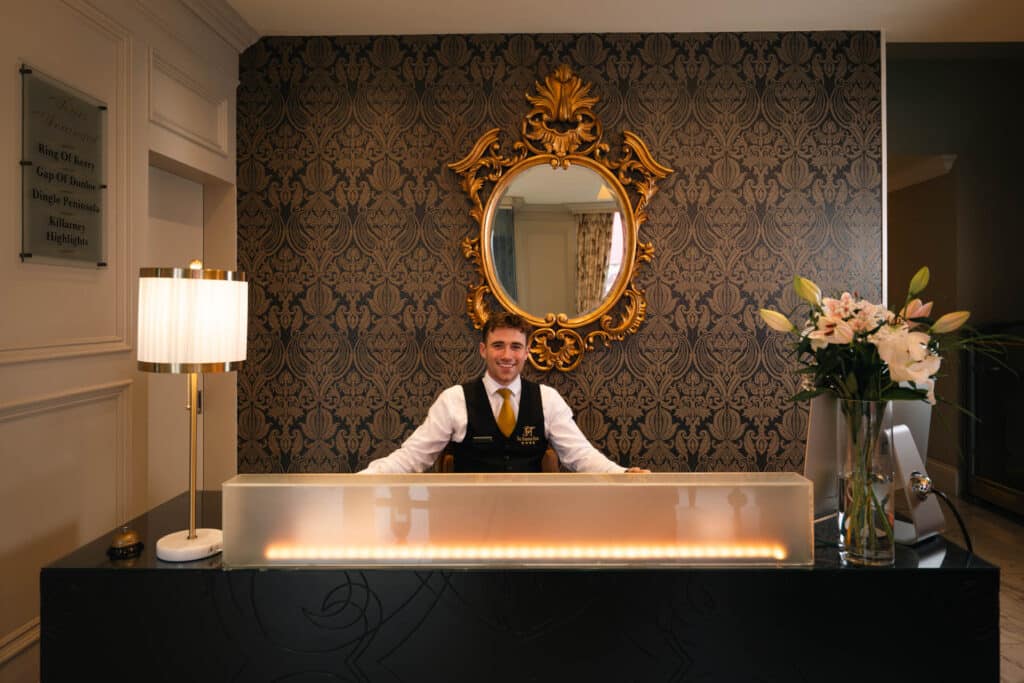 Hotel reception desk with staff member speaking on the phone, decorative mirror and floral arrangement behind the counter.