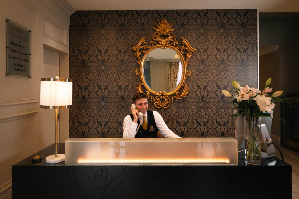 Hotel reception desk with staff member speaking on the phone, decorative mirror and floral arrangement behind the counter.