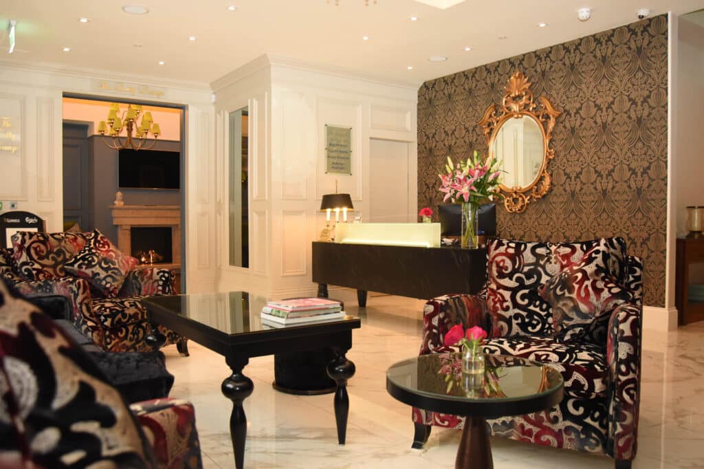 Hotel reception area with front desk, patterned armchairs, decorative mirror and seating tables.
