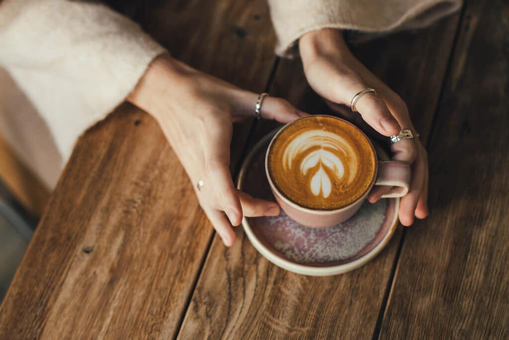 Hands holding a cup of coffee with latte art on a wooden table, creating a warm and relaxed café atmosphere.