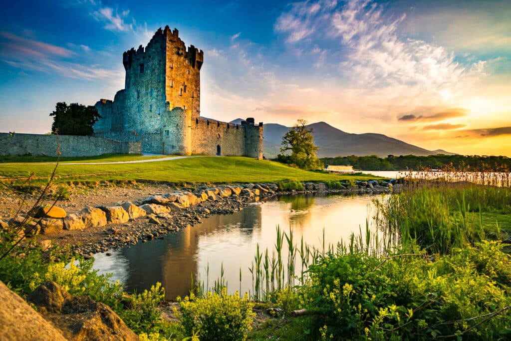 Ross Castle in Killarney during golden hour, overlooking calm water with surrounding greenery and mountains in the background.
