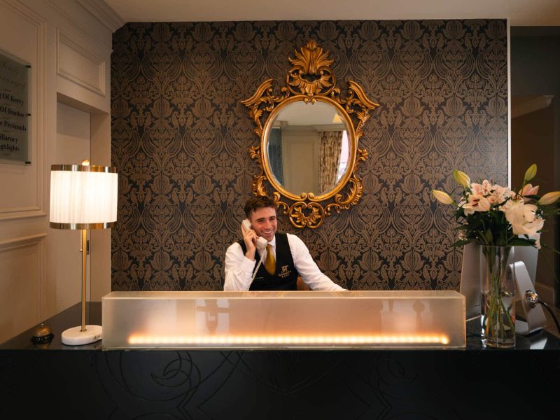 Hotel reception desk with staff member speaking on the phone, decorative mirror and floral arrangement behind the counter.