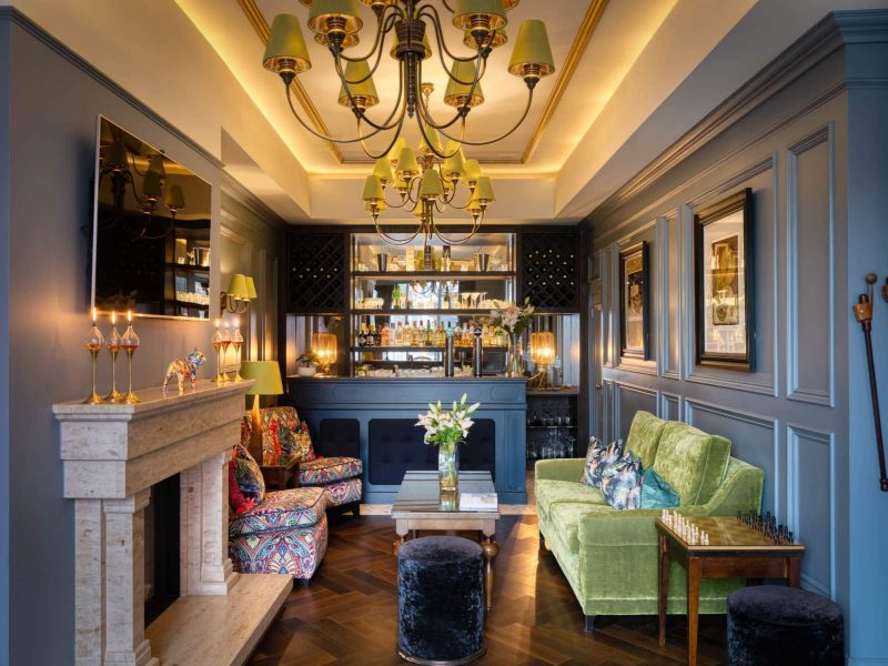 Hotel lounge and bar area with seating, fireplace, chandeliers and drinks counter.